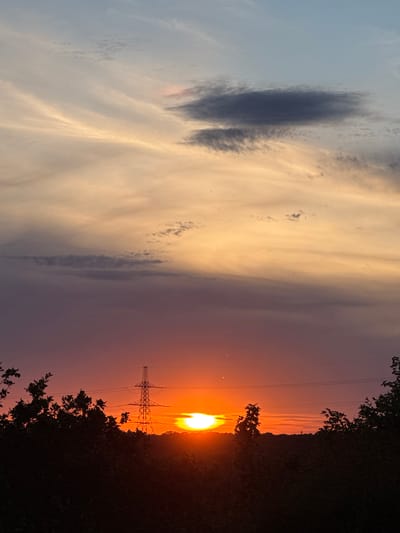 The image depicts a sunset over a landscape, likely Grays, United Kingdom, given the presence of a pylons which are common in the area's industrial and open spaces. The sky displays a gradient of colors, transitioning from a pale blue at the top to warm oranges and reds near the horizon. Wispy clouds are scattered throughout the sky, with one prominent dark gray cloud formation near the top.
In the mid-ground, a tall electricity pylon stands silhouetted against the vibrant sunset. Power lines stretch horizontally from the pylon across the scene. The foreground is dominated by the dark silhouettes of trees and foliage, obscuring the ground below. The sun itself is a bright, intense orb peeking through the horizon, casting a warm glow. The overall impression is of a rural or semi-rural setting at dusk. There are no people or discernible actions depicted in the image. The environment is natural, with man-made structures integrated into the landscape.