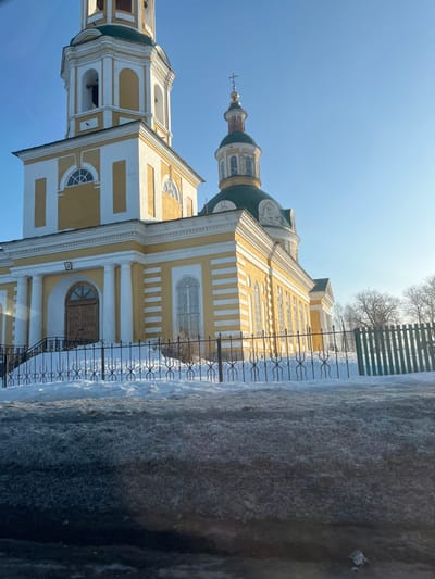 The image depicts the Church of the Assumption of the Blessed Virgin Mary in Чутырь, Russia, under a clear, bright blue winter sky. The church features a dominant yellow facade accented with white architectural details, including pilasters, cornices, and horizontal banding. On the left, a tall bell tower with arched openings and a green roof rises. To its right, the main church body is visible, topped by a large green dome and a smaller golden onion dome crowned with an Orthodox cross. The main entrance, a double wooden door, is approached by steps. The ground around the church is covered in snow. A black wrought-iron fence with decorative scrollwork runs across the mid-ground, separating the church grounds from the foreground, which consists of a dirty, slushy, and icy surface. Bare deciduous trees are visible in the background to the right. No individuals or active subjects are present. The sunlight is strong, causing a lens flare on the left side of the frame.
