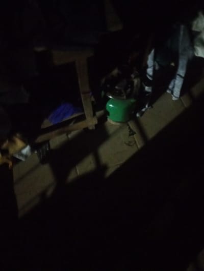 A green gas cylinder is positioned on a wooden shelf in a dimly lit room. The shelf is constructed from rough-hewn wooden planks and is supported by vertical wooden legs. Above and behind the cylinder, several pieces of fabric, appearing to be clothing, are draped over other parts of the shelf. To the left of the gas cylinder, a blue cloth is visible. The floor appears to be made of concrete tiles. The overall scene suggests a storage area or living space with basic furnishings. The presence of the gas cylinder implies its use for cooking or heating.