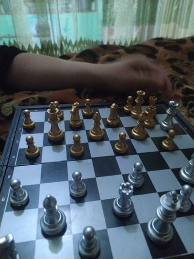 The image displays a close-up, elevated view of an active magnetic chessboard resting on a patterned blanket. The chessboard features alternating light gray and dark gray/black squares. Numerical algebraic notation (ranks 2 through 7) is partially visible along the left edge, indicating a standard board orientation where the 'White' side is toward the viewer.

Two distinct sets of chess pieces are present: one set is silver-colored, and the other is gold-colored.
The silver pieces, positioned in the foreground (White's side), include pawns on a7, c7, e7, and g7. On rank 6, a silver rook is positioned on c6, a silver bishop on d6, a silver pawn on e6, and another silver pawn on f6. Additional silver pieces are partially visible along the bottom edge of the frame (implied rank 8).
The gold pieces, positioned in the background (Black's side), include pawns on a2, b2, c2, d2, f2, g2, and h2. On rank 1 (partially visible at the top edge of the frame), a gold king is on d1, a gold knight on e1, and a gold bishop on f1.

A human forearm, light-skinned and bare, extends horizontally from the left across the upper background portion of the image. The forearm rests on a brown, black, and orange leopard-print patterned blanket or bedspread. Behind this, a blurred light green or teal curtain with a distinct pleated top edge is visible, suggesting an indoor setting, likely a room with a window. The scene is illuminated by ambient light, casting soft shadows beneath the chess pieces. No visual information within the image confirms the specified location as Limbur, Uzbekistan.