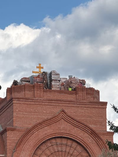 This is a vertical, eye-level, medium shot photograph of the upper portion of a brick church construction in Il’inskiy, Russia. The sky is partly cloudy with patches of blue. A golden Eastern Orthodox cross is positioned atop the building. Several workers are visible on the rooftop, some wearing masks. Building materials, including stacks of bricks and buckets, are scattered around the site. The foreground features a red brick facade with arched architectural details. A pine tree branch is partially visible on the right side of the image.