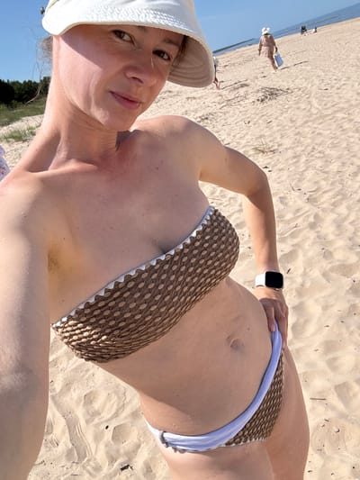 An adult female, wearing a white bucket hat and a brown patterned bikini, stands on a sandy beach. Her left arm is extended, holding a smartphone to take a selfie. The woman has a smartwatch with a white band on her left wrist. In the background, a person in a light-colored outfit walks away from the camera, carrying a blue bag. Other individuals and a stroller are visible further down the beach, near the water. The environment consists of sand, sparse vegetation, and a body of water under a clear sky. This scene is located on the beach in Kalngale, Latvia.
