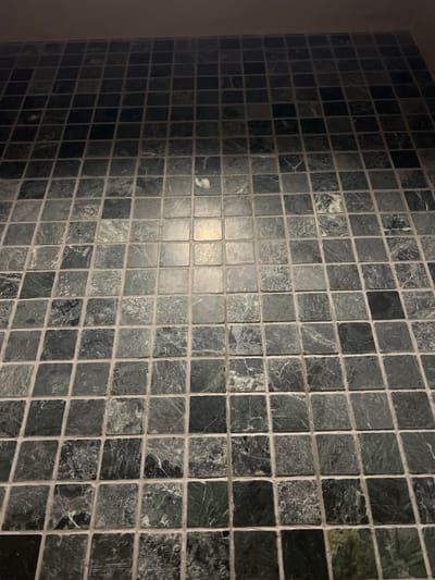 The image shows a close-up view of a tiled surface, likely a floor or wall. The tiles are small, square, and have a marbled pattern with shades of dark green, gray, and white. The grout between the tiles is light-colored. The lighting suggests an indoor environment, possibly with a light source directly above the tiles, creating a bright reflection in a few of the tiles. The angle of the photograph is from a slightly elevated position looking down at the tiled surface. The location of Vyshen'ky, Ukraine is not discernible from the image itself.
