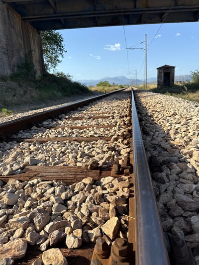 The image shows a close-up, low-angle view of a train track. The perspective is angled, following the line of the rails into the distance. The tracks are made of metal and are supported by wooden sleepers, with gravel ballast filling the space between and around them. The sunlight casts long shadows across the gravel, suggesting it is either early morning or late afternoon.

In the background, a small, weathered stone structure resembling a signal box or a small shed stands by the side of the track. Beyond that, a clear blue sky with a few scattered white clouds stretches over a landscape of rolling hills and distant mountains. Overhead, power lines and poles for an electrified railway are visible. On the left side of the frame, the edge of a concrete structure, possibly a bridge or overpass, is visible, with some greenery and trees growing around it.

The scene appears to be set in a rural or semi-rural area, with the train tracks leading through a natural landscape. There is no visible activity or people. The overall mood is one of quiet solitude and a sense of journey. The location is identified as Mojanovići, Montenegro. No visible text is present in the image.