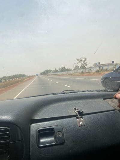 The image depicts a view from inside a vehicle, likely a car or truck, traveling on a highway. The road is paved and has lane markings. The surrounding environment appears dry and dusty, with trees and vegetation visible along the roadside. In the foreground, part of the vehicle's interior is visible, including the dashboard with a latch and a handle, suggesting the presence of a storage compartment. A passing car can be seen to the left. It is challenging to ascertain the precise location as Zangon Aya, Nigeria, with certainty from this image, but the environmental conditions and vegetation are consistent with that region.