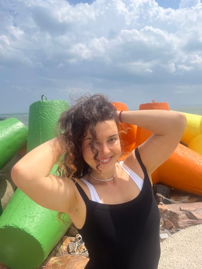 A young woman with dark, curly hair is posing for a photo outdoors. She is wearing a black tank top over a white bra and a beaded necklace. Her hands are raised to her head. In the background, there are large, colorful concrete blocks, painted green, orange, and yellow, positioned along what appears to be a shoreline with a body of water and a hazy sky with clouds. The location is identified as Phú Thủy, Vietnam.