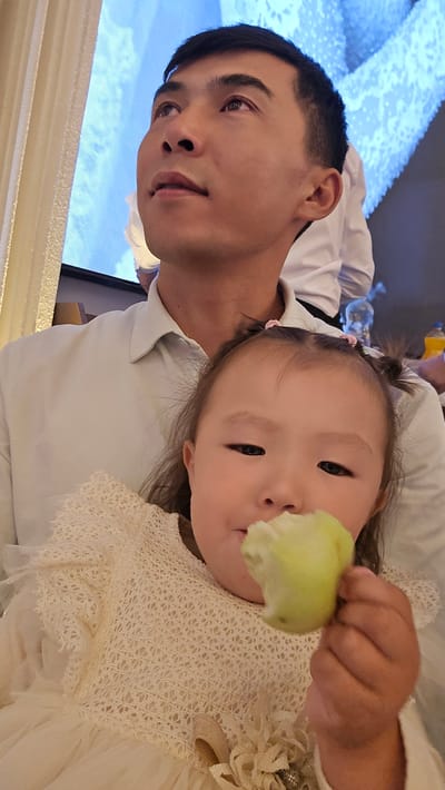 In Karakol City, Kyrgyzstan, a medium shot captures a man and a young girl. The man, positioned behind and to the left, is looking upwards and to the left, his head angled. He has short black hair and appears to be wearing a light-colored collared shirt. He is holding the girl. The girl, in the foreground, is facing the camera and eating a green apple. She has dark hair styled in two pigtails with pink ties and is wearing a white lace dress. The background is blurred, showing what appears to be a large screen displaying a blue and white abstract pattern, and some indistinct figures. The lighting suggests an indoor setting, possibly a restaurant or event space.