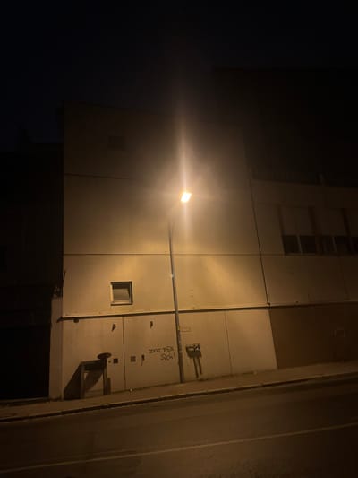 This nighttime photograph captures a stark urban building facade, likely located in Bamberg, Germany, illuminated by a single, prominent street light. The light, positioned towards the center-left, emits a strong, warm yellow-orange glow, creating a significant lens flare and brightly illuminating the light-colored, paneled surface of the building. The surrounding areas, particularly the sky to the left, are cast in deep shadow, emphasizing the artificial light source.
The building's facade is composed of large, rectangular panels, showing seams and subtle textures. Several small, dark, square-shaped openings or windows are visible across the illuminated wall. Above the main section, glimpses of what appear to be upper floors or more windows can be seen in the dim light. A thin pole extends horizontally from the light fixture, supported by another slender bar attached to the wall.
On the lower right section of the illuminated wall, there is dark graffiti. This includes a small, stylized, abstract drawing, possibly resembling a creature or symbol. Below this drawing, the German phrase "Zeit Für Sich!" is spray-painted in dark letters. This translates to "Time for oneself!" or "Time for yourself!". To the far right, a darker, vertical architectural element or an adjacent building wall frames the scene. The overall impression is one of a quiet, artificially lit streetscape after dark.