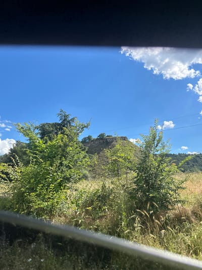 A bright, sunny day is depicted with a clear blue sky and scattered white clouds. In the foreground, lush green bushes and tall grasses dominate the scene, with some dry, golden grass also visible. A dark, horizontal band runs across the bottom of the image, obscuring part of the foreground.
In the middle ground, a gentle, grassy hill rises, topped with what appears to be a small, ancient fortress or ruin. The hill is dotted with sparse trees. Further in the background, a dense line of green trees covers another, steeper hillside. Several thin, dark power lines stretch horizontally across the sky, connecting unseen points.
The location is described as the city of Beloslav, Bulgaria. The scene suggests a rural or semi-rural area with historical remnants. There are no people or animals visible in the image. The overall impression is one of a tranquil, natural landscape bathed in sunlight.