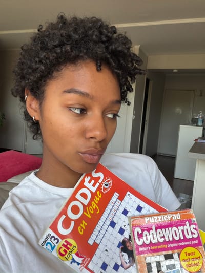 A young woman with dark, curly hair and brown skin is holding two codeword puzzle books. One book is French, titled "Codes en Vogue," and the other is English, titled "Codewords." She is wearing a white t-shirt and appears to be sitting on a pink surface. The background shows a kitchen or similar interior with white cabinets and appliances. The photo was taken inside an apartment in Champs-sur-Marne, France.