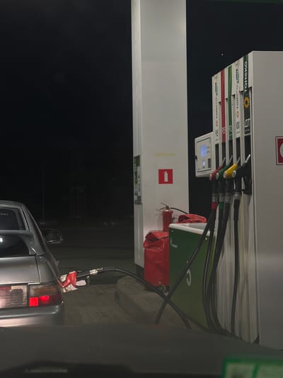 At night, a silver car is positioned at a gas pump at a Tatneft gas station in Tomilino, Russia. The gas pump features multiple fuel dispensers, some labeled with numbers like 95, 92, and 100, and brand names such as "TONECOBIO" and "TAHEKO". A red fuel nozzle is inserted into the car's fuel intake. The scene is illuminated by artificial lighting. In the background, rows of dimly lit fuel dispensers are visible, along with a dark, empty sky. A red fire extinguisher is also present near the pumps.