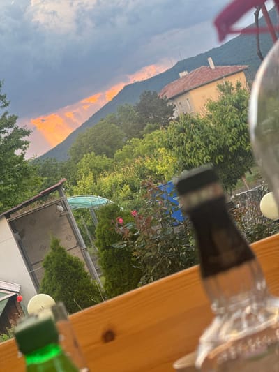 The image shows an outdoor scene, possibly taken from a balcony, in Srednia, Bulgaria. The camera is oriented at an angle, with blurred foreground elements, including a wooden railing and glass objects, partially obscuring the view. In the background, a building with a red roof and yellow facade is visible, nestled against a mountain backdrop. The sky is partially overcast with dark clouds, but a vibrant orange glow is visible between the clouds and the mountain, suggesting either a sunrise or sunset. Lush green trees and vegetation fill the middle ground. There is a wire cage structure with a white object underneath.