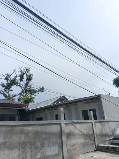 The image displays an overcast daytime scene in what appears to be a residential or developing area, consistent with the provided location context of Carcar, Philippines. The upper two-thirds of the frame are dominated by a pale grey, cloudy sky, through which numerous dark utility and communication lines crisscross diagonally and horizontally, forming a dense network.
Below the power lines, two trees with prominent, somewhat bare branches and green foliage are visible; one on the left and another further right, situated behind the buildings. Small green plants, possibly epiphytes, can be seen clinging to the tree branches.
In the midground, behind a weathered, raw concrete wall with visible vertical support posts, two structures are present. On the left, a partial view of a building with a reddish-brown tiled roof and dark windows or door openings is visible. To its right, a single-story house is under construction or renovation. Its walls are unfinished, unpainted grey concrete or stucco, showing patch marks and raw textures, with several rectangular openings for windows or doors. This house features a grey, corrugated metal roof.
The foreground is occupied by the textured concrete wall, which acts as a barrier. A small, rough patch of ground or debris is visible in the bottom right corner. There is no visible human activity or text in the image. The scene conveys a quiet, perhaps developing, urban landscape under a cloudy sky.