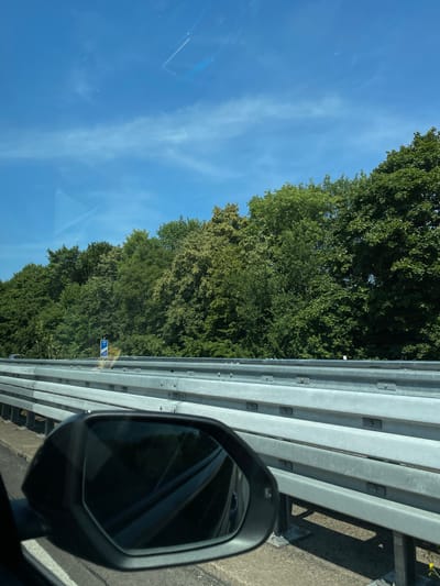 This is a street-level, eye-level, outdoor, daytime photograph taken from a vehicle traveling on a highway in Sankt Augustin, Germany. The foreground shows the driver's side mirror and the car's interior. The middle ground consists of a metal highway guardrail, a concrete barrier, and a blue road sign indicating a speed limit of 100 km/h. Beyond the guardrail is a dense line of green trees that stretches across the horizon. The background is a clear blue sky with faint white contrails. The lighting is bright, indicating a sunny day. The scene depicts a typical highway environment in a suburban or rural area.