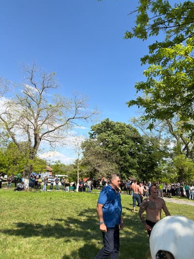 The image depicts an outdoor scene in Dragomir, Bulgaria. A grassy area with scattered trees and a clear blue sky provides the backdrop. A crowd of people stands near what appear to be some temporary structures or food stalls. In the foreground, a man in a blue shirt and dark pants stands facing the crowd. A shirtless man with tattoos on his upper body is positioned near him, partly cut off by the frame, with other shirtless men visible in the background. This suggests some sort of event or competition is taking place. The overall environment appears relaxed and informal.