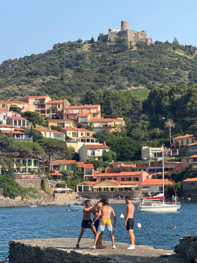 In the city of Collioure, France, a scene unfolds by the clear blue water. On a stone dock in the foreground, three young men are engaging in playful physical interaction. Two of the men, wearing swim trunks, are wrestling or playfully tussling with each other. The third man stands watching them, also in swim trunks. Beyond the dock, a white sailboat with a red sail is moored in the bay. The background is dominated by a hillside densely covered with green trees and dotted with houses with terracotta roofs, characteristic of the Mediterranean architecture. At the very top of the hill, an old stone castle stands as a historical landmark. The sky is a clear, bright blue.