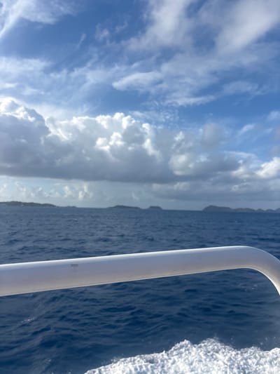 The image displays an open water view from a moving vessel in the vicinity of Spanish Town, British Virgin Islands. A white tubular handrail, consistent with a boat railing, extends horizontally across the lower-mid frame, curving upward on the right side. Below the handrail, a white foamy wake is visible on the dark blue, rippled ocean surface, indicating the vessel's forward motion. On the distant horizon, multiple low-lying landmasses, appearing as islands, are silhouetted against the sky. The sky features a mix of bright white cumuliform clouds and darker grey clouds, with interspersed areas of clear blue sky, particularly in the upper portion. No individuals are depicted within the frame.