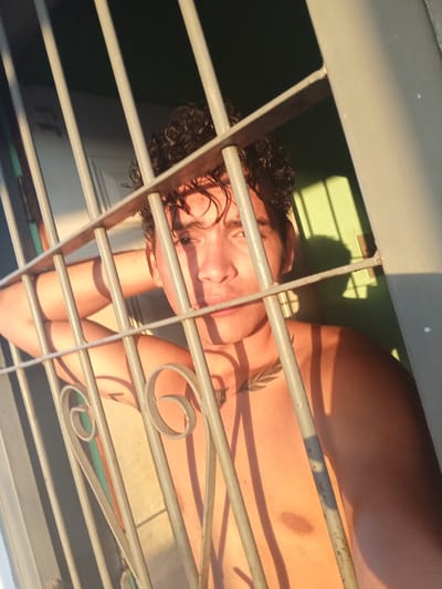 The image shows a close-up, waist-up view of a young man with curly dark hair, leaning against a metal fence. The fence has vertical bars and decorative scrollwork. He is positioned behind the fence, looking towards the camera with his left arm resting behind his head. The lighting suggests it is either sunrise or sunset, with warm light casting shadows across his face and body. He has a tattoo on his chest. The background appears to be a wall, painted in muted tones, with shadows of the fence bars visible on it. The setting is outdoors, possibly a balcony or a window with a protective grille. The image does not contain any specific landmarks or elements that would definitively place it in La Guardia, Venezuela.