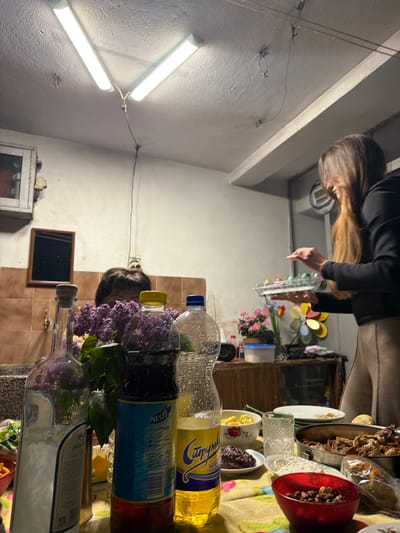 A photograph taken in what appears to be a kitchen or dining room in Damianitsa, Bulgaria. The lighting is provided by two fluorescent lights mounted on the ceiling. A woman is standing near a table, seemingly placing or adjusting food on it. Various dishes and bowls filled with food are arranged on the table, along with beverage bottles, including "Nestea" Lemon flavored. Purple flowers are clustered in bottles. A person's head is visible behind the flowers. A small mirror is mounted on the wall. The environment suggests a homely, indoor setting.