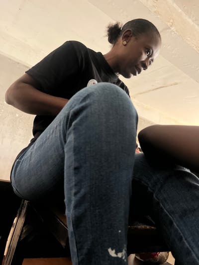 The image is an upward-angled shot of two people, with a focus on the first individual. The first person is a dark-skinned woman with short, dark hair pulled back into a small ponytail. She is wearing a black t-shirt and looking up and to the left. A small stud earring is visible in her left ear. The second person is only partially visible, with their lap and legs prominently featured in the foreground. They are wearing blue jeans. The setting appears to be indoors with off-white walls visible in the background. It is not possible to confirm that the photo was taken in Tudun Nupawa, Nigeria, from the image alone.