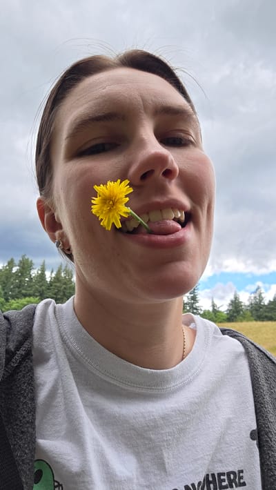 A close-up, waist-up shot features a person holding a yellow dandelion flower in their mouth, their tongue visible between their teeth. The person is wearing a white t-shirt and a gray zip-up jacket. The background consists of a cloudy sky with glimpses of blue sky, green trees, and a grassy field, suggesting an outdoor location. The image is captured in Portland, Oregon, United States of America.