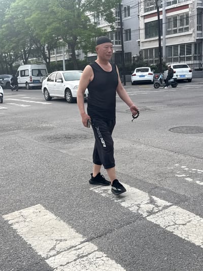 A man is standing in a crosswalk on a street in Linghe, China. He is wearing a black tank top, black cropped pants with "PEAK BOXER" printed on the leg, black sneakers, and a black cap. He is holding a beaded bracelet in his right hand and a cell phone in his left hand. The street has a number of parked cars, a scooter, and some buildings. Trees are visible in the background.