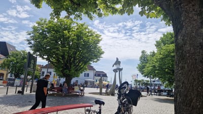 This is a photograph of a public square in Überlingen, Germany, likely near the harbor given the presence of a large white boat in the background. The scene is set on a sunny day with scattered white clouds in a blue sky. Large trees with lush green foliage are prominent in the foreground and midground, with one particularly large tree trunk on the right side of the frame.
In the midground, a statue featuring a person on horseback atop a pedestal is visible. To the left of the statue, a building with white walls and blue shutters stands. A group of people are seated on a red bench in front of this building, appearing to be resting or socializing. A man in a black shirt and pants is walking away from the viewer towards the right side of the frame, his hands in his pockets.
In the foreground, a red bench is positioned partially in view, and a bicycle with a black basket is parked. The ground is paved with light-colored cobblestones. Further in the background, there are more trees, flags, and a harbor with a boat. Other people are scattered throughout the square, some appearing to be walking or standing. A large digital display board is visible on the left side of the image. The overall atmosphere appears to be relaxed and public.