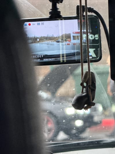 The image depicts a view from inside a vehicle, looking outwards through a dusty or moisture-speckled windshield. In the foreground, a black smartphone is mounted vertically, actively recording video. A red recording dot is visible on its screen, along with several icons typical of a camera interface. The phone screen displays the date "2026/04/21" and a timestamp "01:27", likely indicating the video duration rather than the time of day, given the daylight outside. The text "BCDEF" is also visible at the bottom right of the screen, and "Bol" (possibly part of "Bolt" or a similar word) at the top.
A small, dark, stylized object resembling a frog or toad figurine is suspended by a string or wire in front of the windshield, positioned between the smartphone and the glass. Two taut, parallel wires or strings run horizontally across the frame, passing in front of both the phone and the figurine.
The phone's screen reflects the external landscape, revealing a scene with bare trees, possibly snow or ice on the ground, and some distant structures featuring red and white striped elements. This landscape suggests a rural or semi-rural setting in Klenovskoe, Russia, consistent with early spring conditions where remnants of winter might still be present. The light appears diffused, possibly indicating an overcast day. Parts of the vehicle's dark interior frame the top and bottom of the shot.