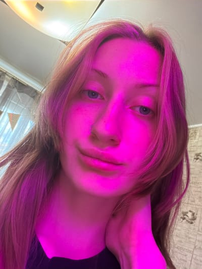 A close-up, head and shoulders, selfie-style photograph taken indoors. A young female with long, reddish-brown hair is positioned in the center of the frame, illuminated by bright pink lighting. Her left hand is raised and resting on her chin, obscuring part of her face. She is looking directly at the camera. A silver nose ring is visible on her left nostril. The background is out of focus and appears to be a room with patterned wallpaper and a curtained window. The image was taken in the city of Novoe Devyatkino, Russia.