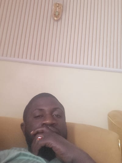 This image depicts an adult male indoors in Babawa, Nigeria. The man, with dark skin, short dark hair, and a beard, occupies the lower half of the frame, looking directly at the camera. He is wearing a light-colored shirt, possibly green or teal. His right hand is raised, with fingers partially obscuring his mouth and nose. A ring with a reddish-brown stone is visible on his right ring finger. He is seated on a light brown or tan upholstered armchair, partially visible on the right side of the frame. The background features a plain light-colored wall in the lower section, transitioning to a ceiling with light-colored vertical paneling in the upper section. An oblong, gold-toned wall sconce is mounted on the ceiling paneling in the upper center of the image.