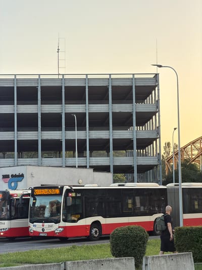 The image shows a street scene in Gdańsk, Poland. The main subjects are two buses, one in the foreground and another partially visible behind it. Both buses are white and red and appear to be public transportation. The bus in the foreground has "2100DJAZD ZA 20 MIN" displayed on its front, indicating a travel time of 20 minutes. Other text visible on the bus includes "2661" and a license plate "GD 860MN". A person with a shaved head, wearing a black t-shirt and shorts, is walking away from the foreground bus, carrying a green backpack.

The setting includes a multi-story parking garage made of metal and concrete. A street lamp is visible extending from the side of the parking garage. Green bushes and grass are present along the side of the road. In the background, a section of a yellow bridge structure can be seen, along with trees. The sky is a light, yellowish hue, suggesting either sunrise or sunset. The overall impression is of an urban environment on a clear day. Visible text also includes "ZHM Gdańsk" and "ZMIANA ZA".