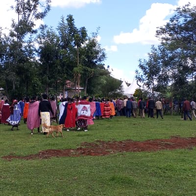 The image shows a group of people in Keyian ward, Kenya gathered on a grassy field with trees in the background under a partly cloudy sky. Some people are wearing traditional Maasai clothing with bright colors and patterns. A dog walks among the group. In the distance, there are white tents set up. A strip of disturbed earth is visible in the foreground.