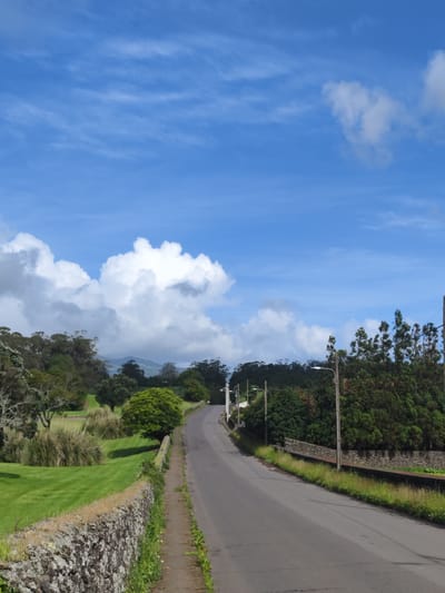 The image shows a paved road curving slightly to the left, lined on either side by stone walls. To the left of the road is a grassy field with some tall grasses and trees in the background. To the right is denser foliage. Overhead are utility poles with wires. The sky is blue with scattered white clouds. The location is in Ribeira Grande, Portugal, as implied by the information provided. No people are visible.