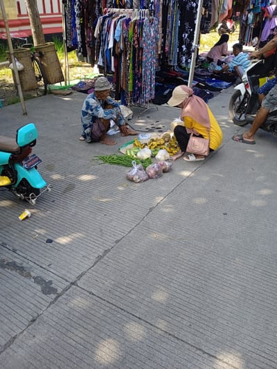 This image depicts an outdoor scene in Kendal, Indonesia. An elderly man with a bucket hat and patterned clothing sits cross-legged on the concrete pavement, barefoot. Next to him, a woman in a yellow shirt, black pants, and a beige sun hat with a pink scarf is squatting. Between them is a collection of produce including bananas, green beans, onions, and garlic, packaged in plastic bags.
Behind them, a clothing rack displays a variety of garments on hangers. Further back, more people are present, some seated on the ground, seemingly also engaged in commerce. A motorcycle is visible on the right side of the frame, with a man in sandals standing nearby.
To the left, a light blue moped is parked, while a bicycle with woven baskets attached to the back is adjacent to a tree. The pavement shows visible grooves and cracks, and the scene is bathed in bright sunlight, casting shadows on the ground.