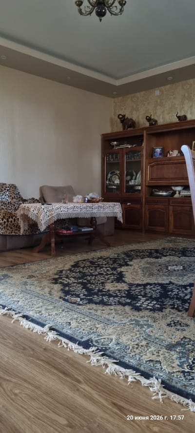The image depicts the interior of a room, likely a living area, with elements suggesting a residence in Alashkert, Armenia. In the foreground, a large, ornate rug with a blue and beige floral pattern covers the wooden floor. To the left, a leopard-print armchair and a beige sofa are partially visible, with a table placed in front of them. The table is covered with a white lace tablecloth and holds several items, including a bowl of fruit, a small box, and some bagged items.

Further back, a tall wooden cabinet with glass doors stands against the wall, displaying various decorative items such as plates and framed photographs. Several elephant figurines are placed on top of the cabinet. The wall behind the cabinet is covered with patterned wallpaper, and a chandelier hangs from the ceiling. The overall impression is that of a furnished, lived-in space. The presence of the date stamp "20 июня 2026 г. 17:57" in the lower right corner is a metadata element from the photograph itself, indicating when the picture was taken.