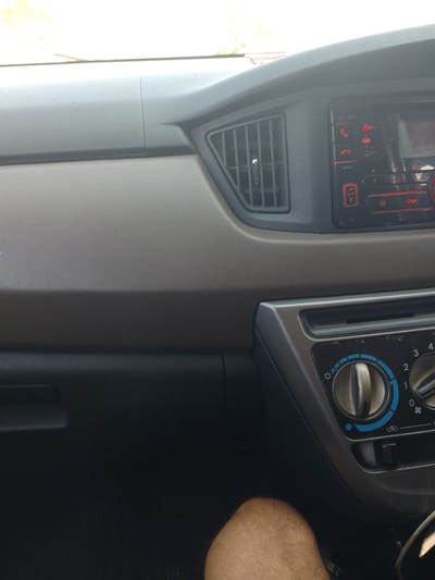 The image shows the interior of a car, focusing on the dashboard and a person's leg. The dashboard features an air vent, a car stereo with a red display and buttons, and climate control knobs. The person's knee and thigh, covered in light-colored hair, are visible in the lower right portion of the frame. The environment appears to be a typical car interior, with textured plastic surfaces. No specific location within Tiang Tara, Indonesia, can be identified from the visual information presented in the image.