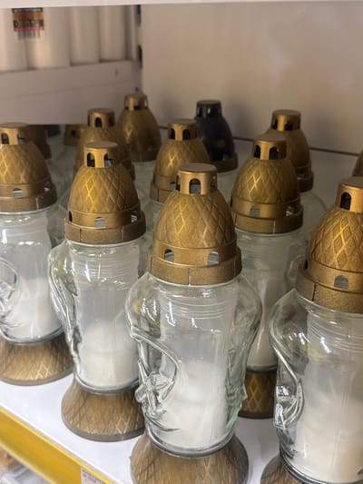 The image displays a close-up, vertical view of several glass memorial candles with decorative gold lids. The candles are arranged in rows on a shelf, suggesting they are in a store or storage area. The lighting appears to be artificial, typical of an indoor setting. The focus is on the objects themselves, with no people visible. The background is a plain, light-colored surface, possibly a wall or another shelf, with some white rolls of material in the far left. A yellow price tag or sign is partially visible on the right side. The candles are the primary subjects, and the scene is static, with no activity taking place. The image is taken from a slightly downward angle, looking across the arrangement of candles. No specific time of day or weather conditions can be inferred. The location context indicates Ivanivka, Ukraine, but there are no specific visual cues within the image to confirm this. There is no discernible text in the image apart from the faint markings on the yellow strip on the right.