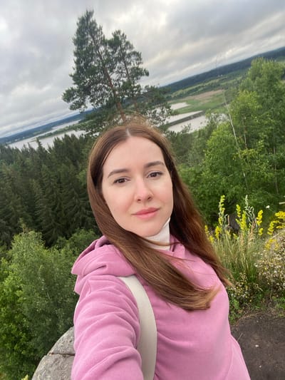 A young woman with long, brown hair is taking a selfie outdoors. She is wearing a pink hooded sweatshirt over a white turtleneck. Her face is turned towards the camera, and she has a slight smile. The background is a lush green landscape with trees, a body of water, and a cloudy sky. The photo is taken from a slightly high angle, looking down at the woman and the scenery. The overall impression is one of a casual, scenic moment. The image appears to be taken during the daytime, with overcast weather contributing to the soft lighting. The location cues suggest a natural environment, possibly a park or a viewpoint overlooking a lake or river.