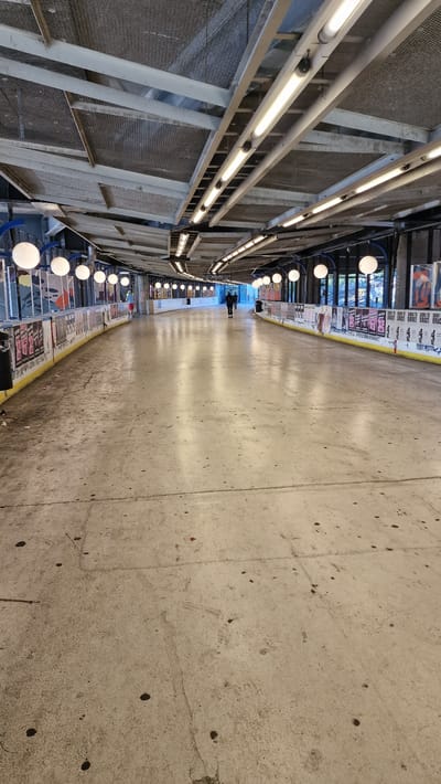 The image depicts an indoor roller skating rink. The rink has a concrete floor with visible cracks and dark spots, and is bordered by a yellow and white barrier with posters attached. The ceiling is constructed with metal beams and mesh panels, with fluorescent lights and circular white lights suspended above. Two figures, appearing to be walking, are visible in the distance on the rink. The environment appears to be a covered walkway or an enclosed space with an open side, suggested by the presence of windows and the structure of the building. The scene is likely a public space designed for recreation.