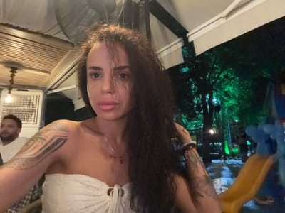 In the city of Petrich, Bulgaria, a woman with dark, curly hair and a white strapless top is shown in a selfie. She has tattoos on her arms and is wearing a necklace with a red pendant. In the background, a man with a beard is visible, and beyond him, the scene transitions to a dark, outdoor environment illuminated by green lights, suggesting a park or outdoor venue with trees and possibly a playground structure.