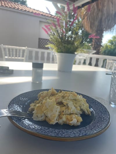 A close-up shot captures a plate of scrambled eggs on a textured blue plate with a dark rim, set on a white table. A fork rests on the left side of the plate. In the background, a salt shaker with a black lid is visible on the table. Beyond that, a blurred potted plant with pink flowers and a palm tree are present, suggesting an outdoor setting. White railing and a tiled roof are also visible, hinting at a building or structure. The environment appears to be a sunny outdoor dining area, possibly at a cafe or restaurant in Skala Avdiron, Greece, characterized by its Mediterranean climate and coastal proximity.