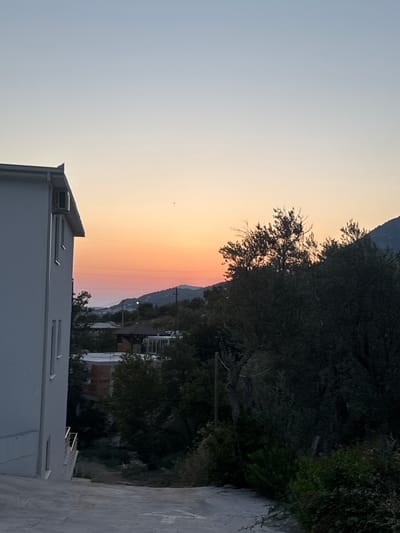 The image depicts a scene in Sustaš, Montenegro, during sunset. A white, multi-story building is prominent on the left, with visible windows and an air conditioning unit. A sloped, paved path leads away from the building. In the middle ground, there are trees, some with dark foliage, and a structure with a brown, peaked roof. Beyond that, rolling hills are visible against a sky transitioning from pale blue at the top to orange and pink near the horizon. The sun is setting behind the hills, casting a warm glow. The overall impression is of a quiet, rural or semi-rural residential area at dusk.