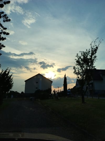 This is a vertically oriented, low-angle photograph taken at dusk in Dobev, Czechia. The scene captures the transition from day to night, with the sun low in the sky and casting a warm glow through scattered clouds. Silhouetted against the bright sky are the upper portions of buildings and trees. The sun's rays are partially obscured by a distinct, wispy cloud formation.

In the foreground, dark silhouettes of evergreen trees are visible along the bottom edge of the frame. A large building with a pitched roof, appearing to be residential or an apartment complex, dominates the middle ground, with its upper floors and roofline silhouetted. To its left, a mature deciduous tree with bare branches is also silhouetted. Further back, the outlines of other structures and trees are discernible, along with a dark grassy area. A narrow, paved path or driveway is visible in front of the building. A thin, dark line along the right edge of the frame may indicate the edge of a vehicle or a railing.

The overall mood is peaceful and serene, characteristic of twilight. The lighting suggests a clear, albeit cloudy, evening. No people or specific activities are visible. There is no visible text in the image.