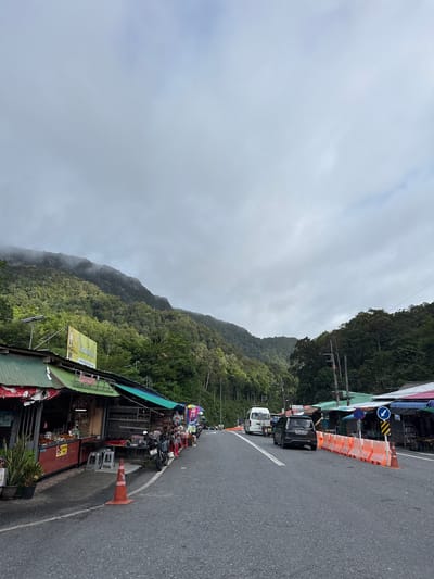The image depicts a roadside scene in Tambon Wang Prachan, Thailand, with a mountain covered in lush green trees and mist in the background. On the left side of the road, there are several small market stalls with awnings, displaying various goods and some potted plants. A motorcycle is parked in front of one of the stalls. An orange traffic cone is placed on the road in front of the stalls.
The road itself is paved and appears to be a two-lane road. Several vehicles are visible, including a dark-colored car in the foreground, a white van further down the road, and a tuk-tuk or similar three-wheeled vehicle. On the right side of the road, there are more market stalls with colorful awnings, and a line of orange temporary traffic barriers separates the stalls from the road. Traffic signs are also visible, including a blue circular sign with a white arrow indicating a turn. The sky is overcast with clouds.