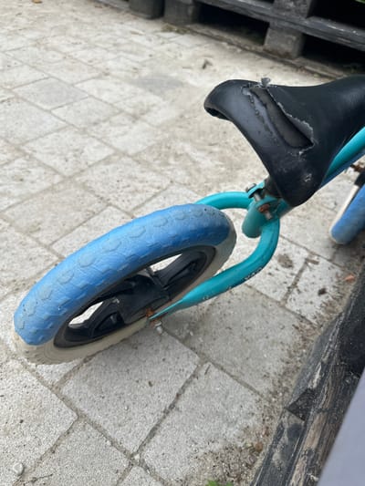The image shows a close-up view of a blue balance bike with a worn black seat, resting on a light-colored paved area. The bike's front tire is blue with a white rim. The background features a dark wooden structure, possibly a fence or bench, and some greenery. The scene appears to be outdoors, possibly in a residential area or park, in the city of Malinovka, Russia.