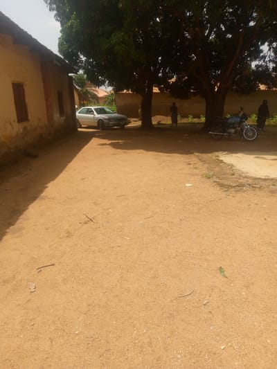 The image depicts an outdoor scene in a village, likely in Madakiya, Nigeria. The foreground is a wide expanse of dry, sandy ground, with scattered pebbles and small sticks. A prominent shadow stretches across the ground from the left, indicating the presence of a building or structure casting it.
To the left of the frame is a rustic building with a weathered, beige-colored wall and a dark, thatched roof. It has a small, dark window. Adjacent to this building, a silver sedan is parked, facing away from the viewer and slightly towards the right. The car appears to be in decent condition.
In the middle ground, several large trees with dense green foliage provide shade. Beneath the trees, a blue and silver motorcycle is parked. Two people are standing under the trees. One, dressed in dark clothing, is partially obscured by a tree trunk and appears to be facing the motorcycle. Another person, also in dark attire, stands further to the right, near the motorcycle.
In the background, a low brick wall runs horizontally, separating the area with the trees and buildings from what appears to be more structures with terracotta-colored roofs. The sky is bright and appears to be sunny, suggesting it is daytime. The overall impression is one of a typical village setting, likely in a warm climate, with vehicles and everyday life activities suggested by the presence of people and parked transport. There is no visible text in the image.