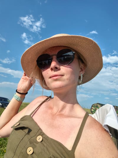 A woman wearing a straw hat and sunglasses is taking a selfie outdoors. She is wearing a green sleeveless dress with wooden buttons and pearl hoop earrings. Her left arm is raised, holding the brim of her hat. A watch with a light blue band is visible on her left wrist. In the background, a dark car is parked in a grassy field under a bright blue sky with scattered clouds. There is also a camouflage chair with a white cloth draped over it visible on the right side of the image. The scene appears to be set in an open, rural area, possibly near Кузьминовка, Russia, given the context. The overall impression is of a sunny day spent outdoors.