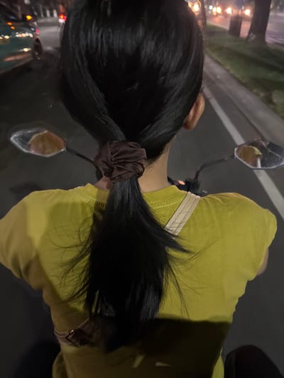 This media file shows a close-up, overhead view of a person's head and shoulders from behind. The subject has long, dark hair that is tied back in a ponytail with a dark brown scrunchie. They are wearing a bright lime green t-shirt with a subtle beige stripe detail near the neckline.
The scene appears to be outdoors, possibly on a street or road, indicated by the presence of a motorcycle mirror reflecting some lights. The background suggests an urban or semi-urban environment at night or dusk, with blurred lights and a paved surface. There are no visible emotions. The location context is South Tangerang, Indonesia.