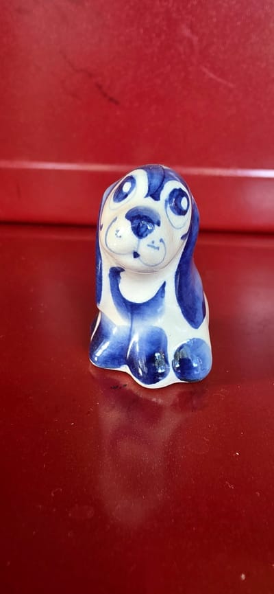 A small, ceramic figurine of a dog, painted with blue and white Gzhel-style patterns, sits on a dark red, glossy surface. The dog figurine is depicted in a sitting pose, with large, expressive eyes and floppy ears. The surface it rests on reflects the figurine and the red background, which appears to be a wall or a piece of furniture. The scene is dimly lit, with the primary light source coming from the front, highlighting the glossy texture of the red surface and casting a soft shadow behind the figurine. The overall impression is one of a simple, decorative object placed on a plain background.