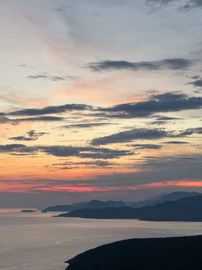 This image depicts a sunset over a body of water, likely a lake or bay, with mountainous terrain in the background. The sky is filled with dramatic clouds, illuminated by the setting sun, creating a vibrant display of orange, pink, and gray hues. The mountains are silhouetted against the colorful sky, appearing as dark, layered shapes. The water reflects the colors of the sky, creating a shimmering effect. On the right side of the image, a dark mass suggests a shoreline or a densely forested area. In the distance, a very small, dark object can be seen on the water, possibly a boat or a buoy.

The time of day is clearly sunset, indicated by the colors in the sky and the low angle of the sun. The weather appears to be calm, with the water being relatively smooth. The presence of mountains and a large body of water suggests a natural landscape, possibly a national park or a scenic coastal region. The overall mood conveyed by the image is one of awe and tranquility, with the dramatic sunset evoking a sense of natural beauty and peacefulness. There is no visible text in the image.